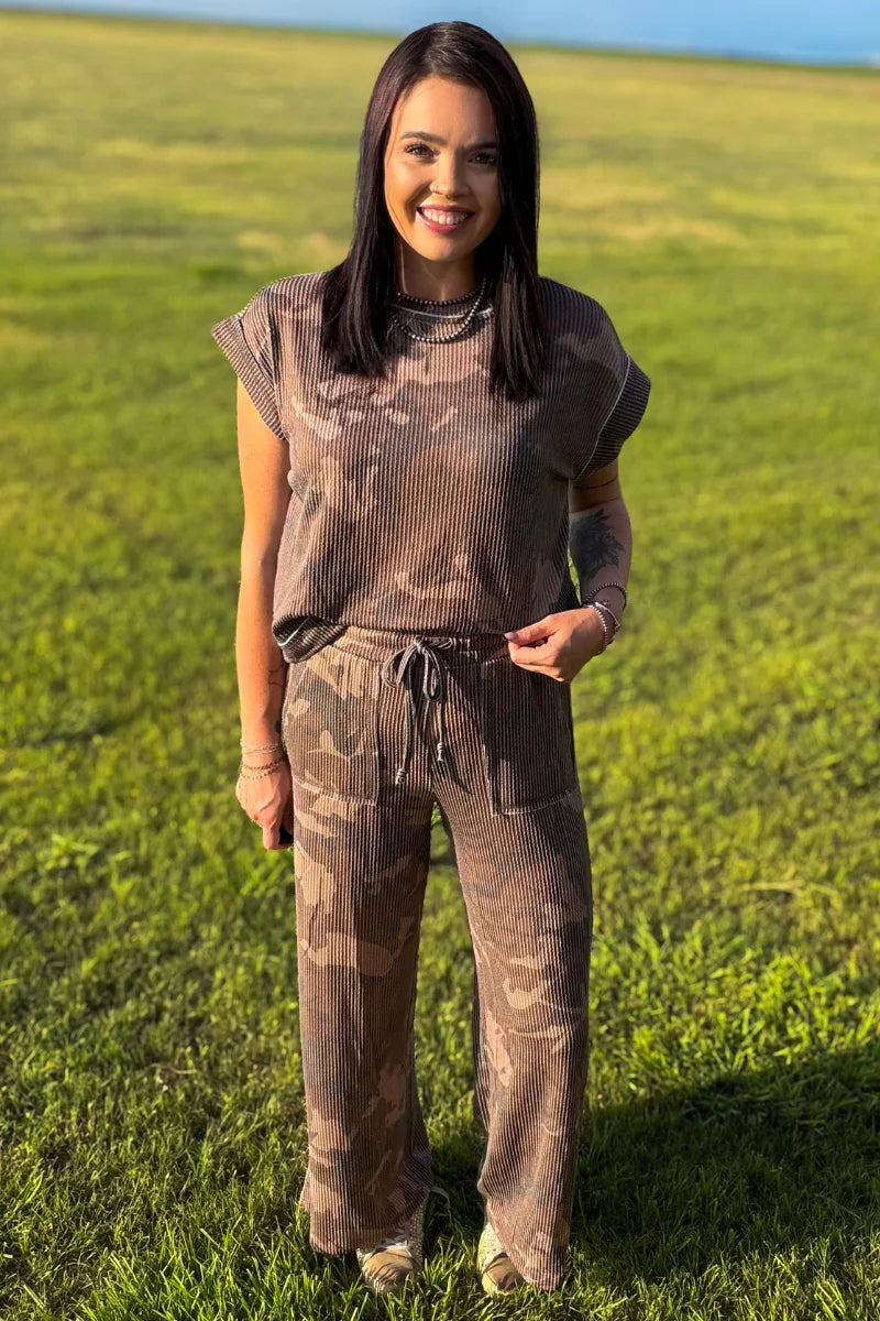 A woman wearing a matching brown camouflage ribbed two-piece set consisting of a short-sleeve top and wide-leg pants, standing in a grassy field.