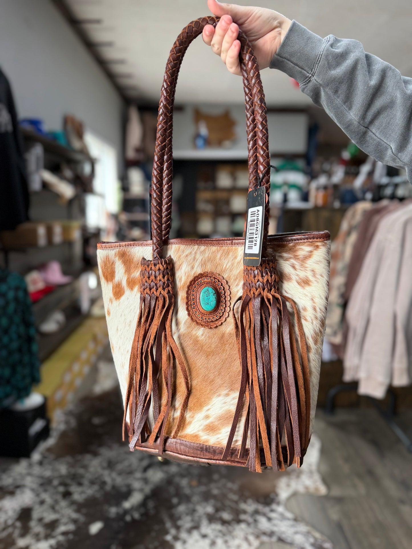 Brown leather handbag with fringe and a turquoise stone held by a person in a store setting.