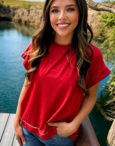 A woman wearing a bright red short-sleeve t-shirt with white contrast stitching, standing outdoors near a body of water.