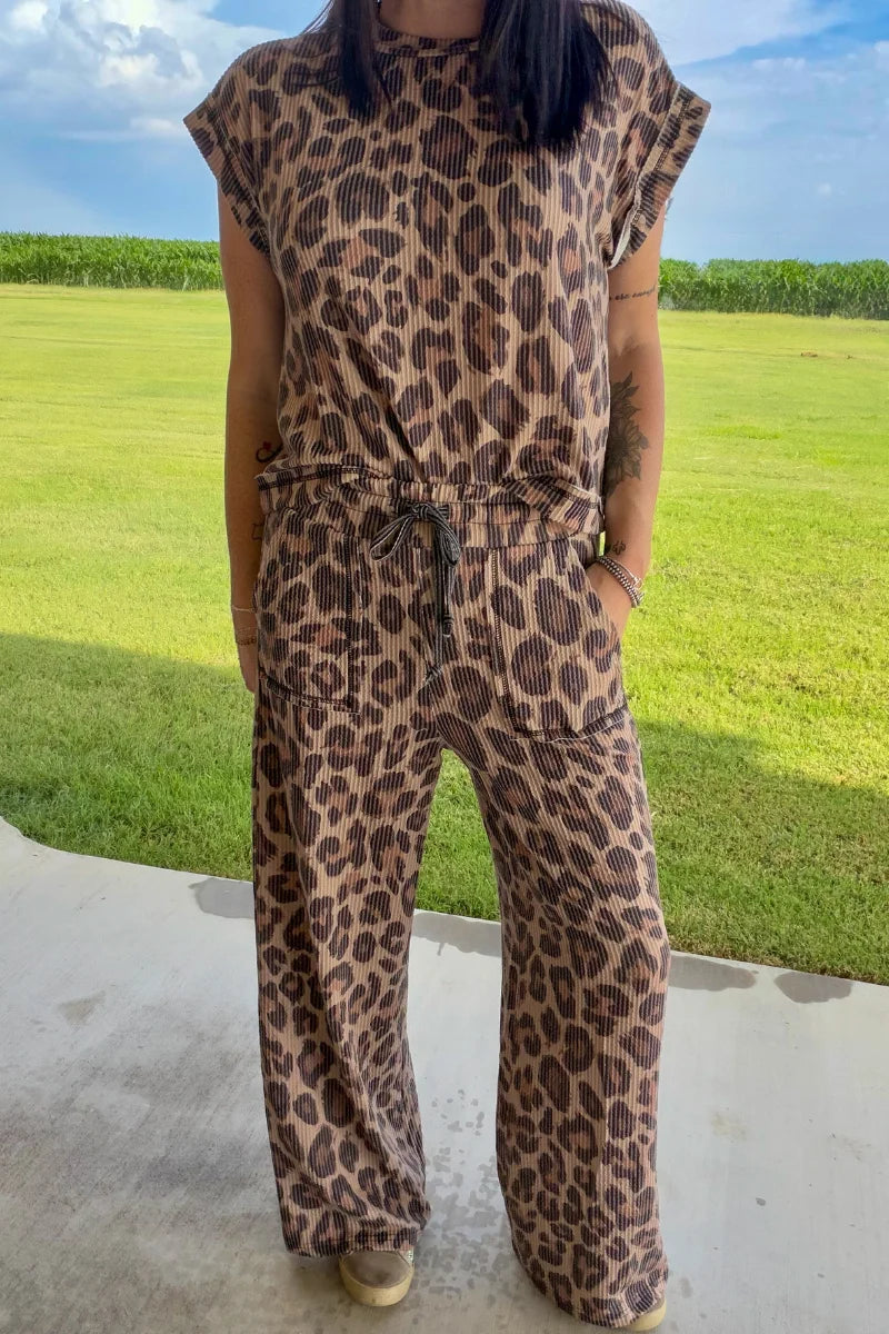 A woman wearing a brown and beige leopard print jumpsuit, standing outdoors on a concrete patio with a green grass field and blue sky in the background.
