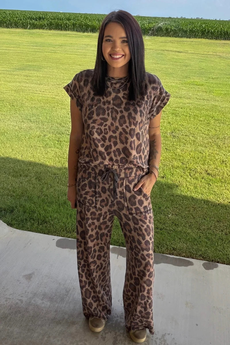 A woman wearing a brown and black leopard print short-sleeve jumpsuit, standing outdoors on a concrete patio with a green grass lawn and cornfield in the background.