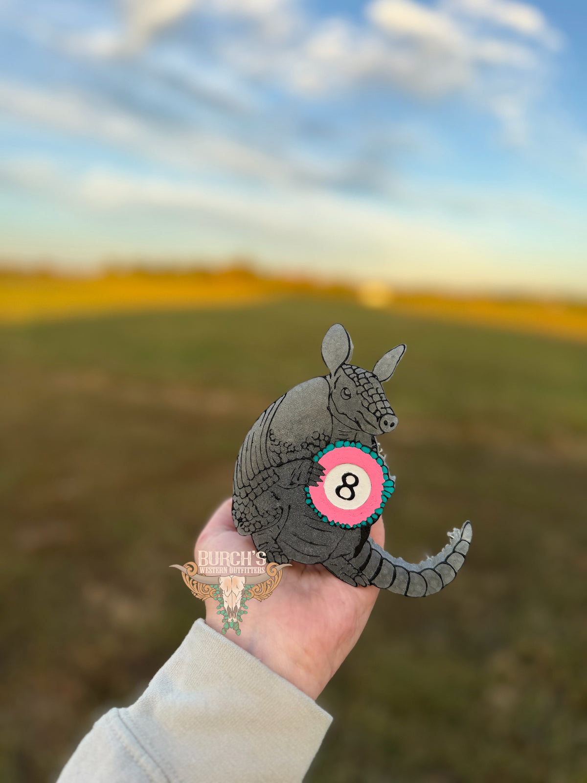 A hand holds a small gray armadillo-shaped item with a pink circular badge featuring the number 8, against a blurred outdoor field and sky background.