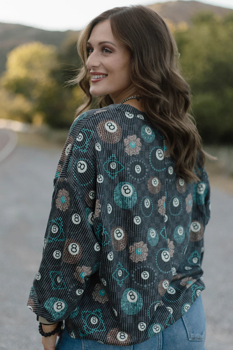 A woman wearing a long-sleeve button-up blouse with a dark teal, black, and white geometric floral pattern, photographed outdoors against a blurred background of trees and hills.