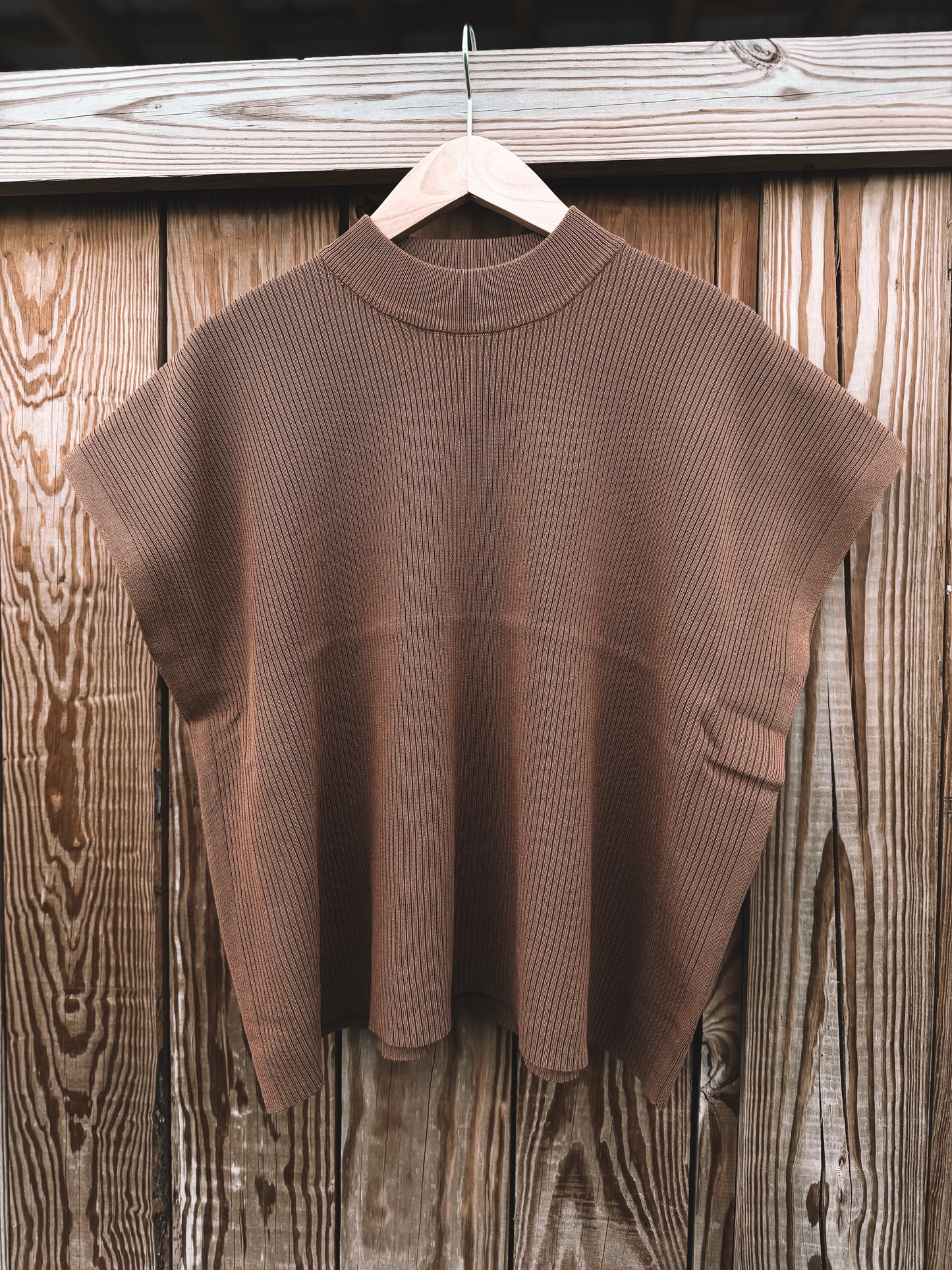 A brown ribbed knit short-sleeve top hanging on a wooden hanger against a rustic wooden plank wall.
