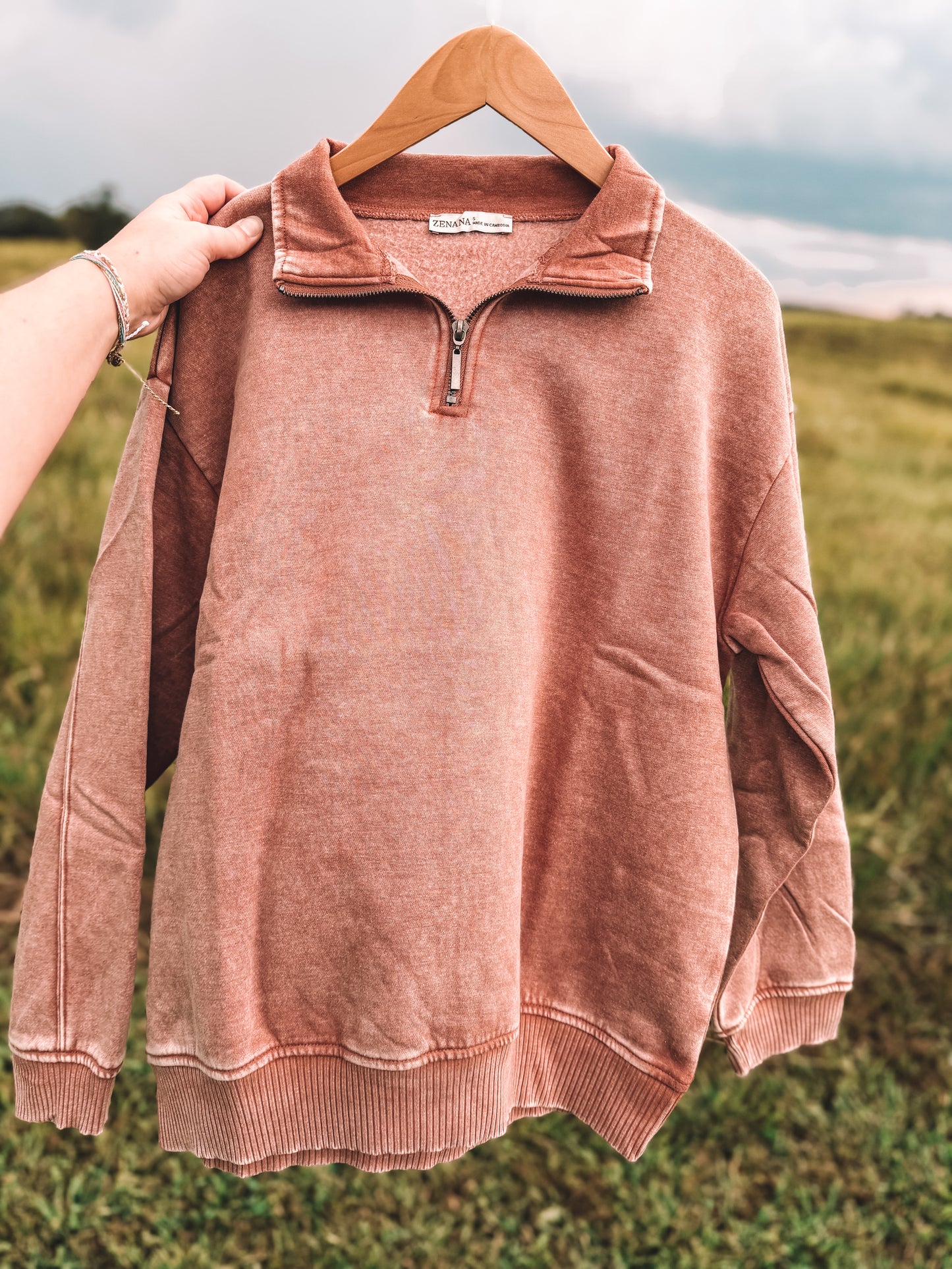 A dusty rose quarter-zip sweatshirt with a ribbed collar, cuffs, and hem, hanging on a wooden hanger outdoors with a hand holding the collar on the left.