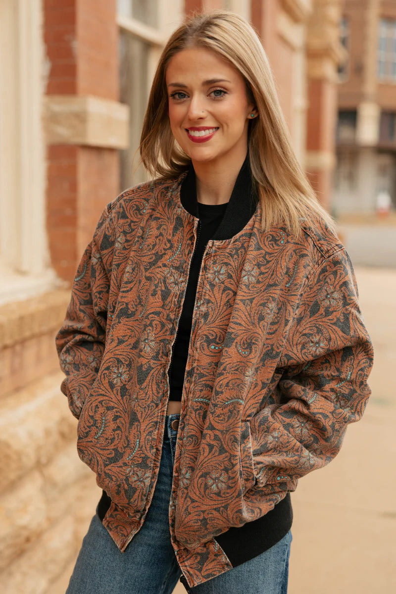 A woman wearing a brown and black paisley patterned bomber jacket over a black top and blue jeans, standing outdoors in front of a brick building.