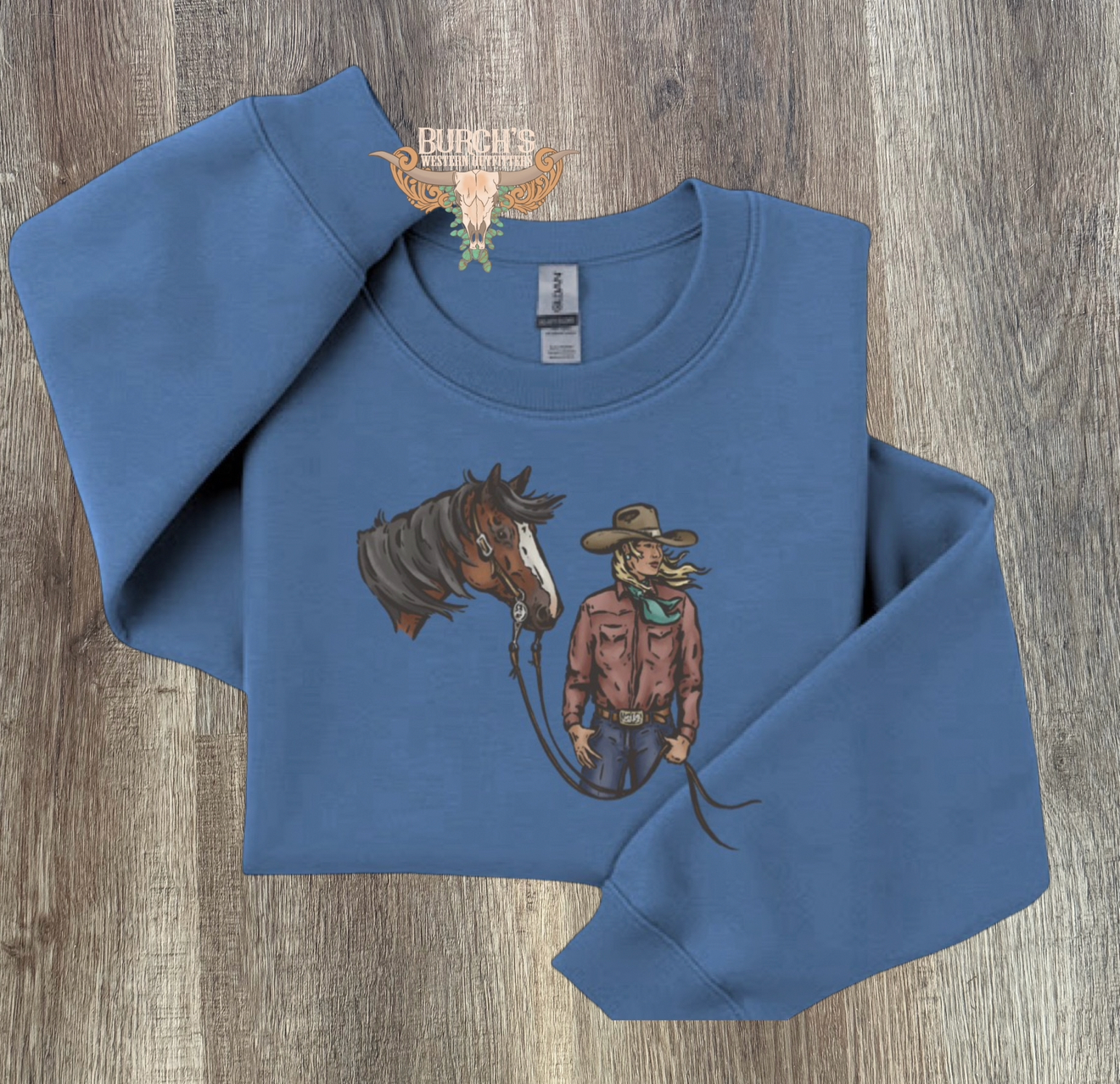 A blue crewneck sweatshirt laid flat on a wood background, featuring a graphic print of a cowboy and his horse.