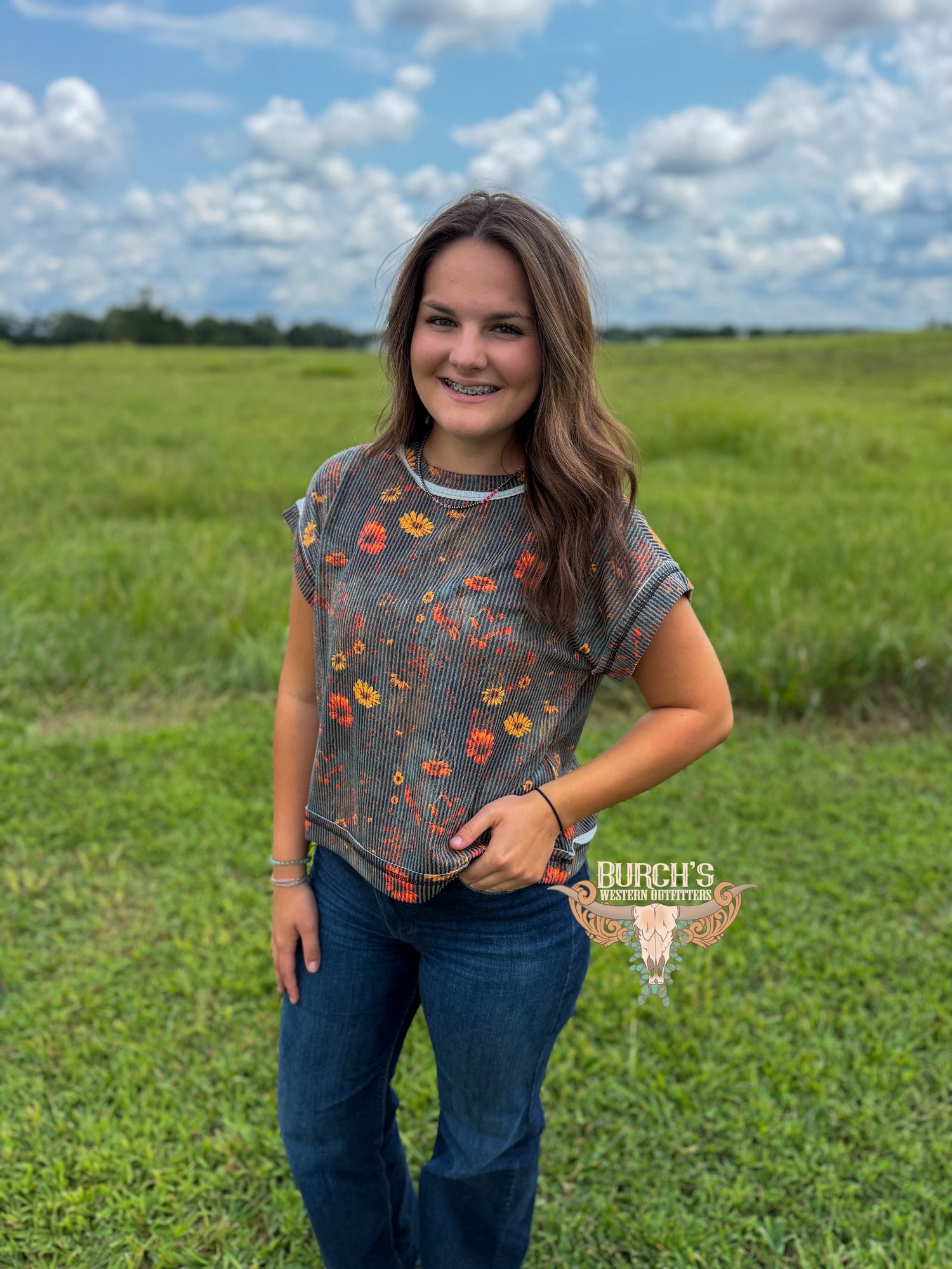 A woman stands in a grassy field under a cloudy blue sky, wearing a short-sleeve gray ribbed top with a colorful floral print and dark blue jeans. A watermark for 'Rurch's Texas Apparel' is visible on the lower right.