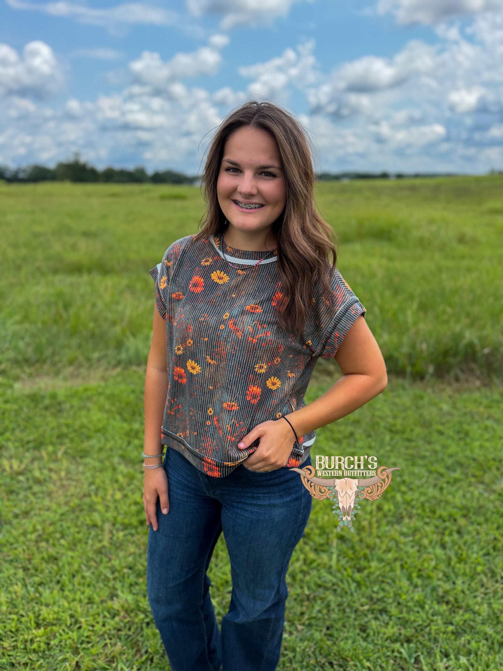 A woman stands in a grassy field under a cloudy blue sky, wearing a short-sleeve gray ribbed top with a colorful floral print and dark blue jeans. A watermark for 'Rurch's Texas Apparel' is visible on the lower right.