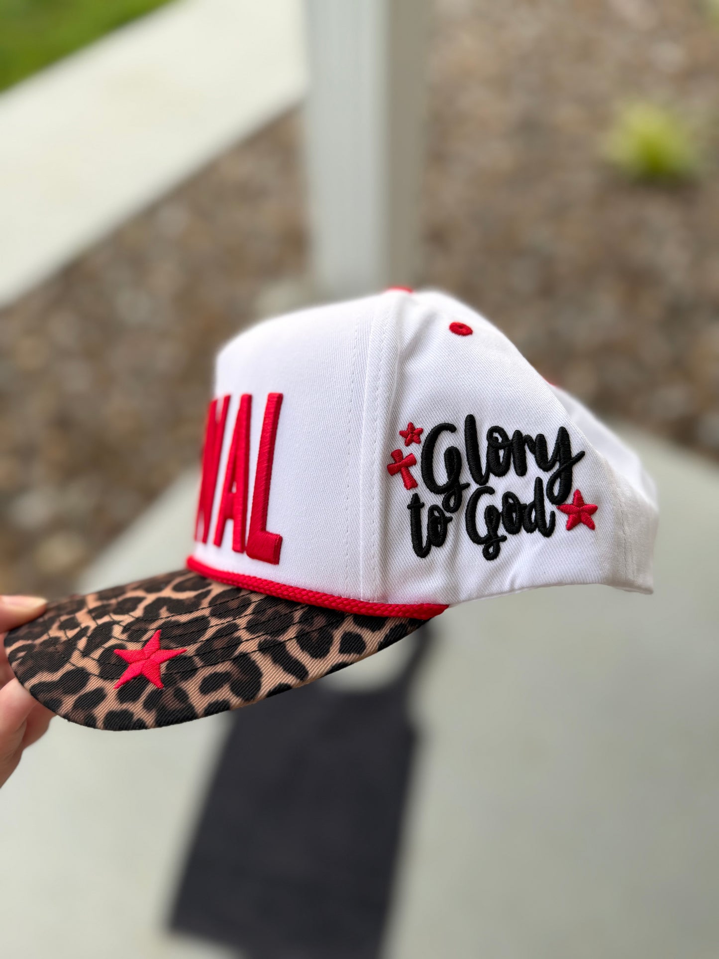 White cap with 'Glory to God' text and leopard print brim held by a hand outdoors.