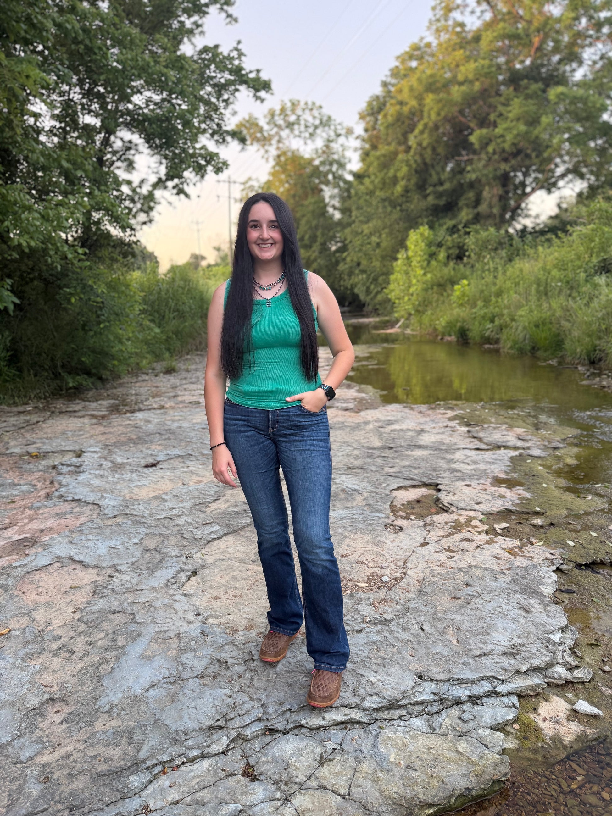 Woman standing on a rocky path by a river with trees in the background