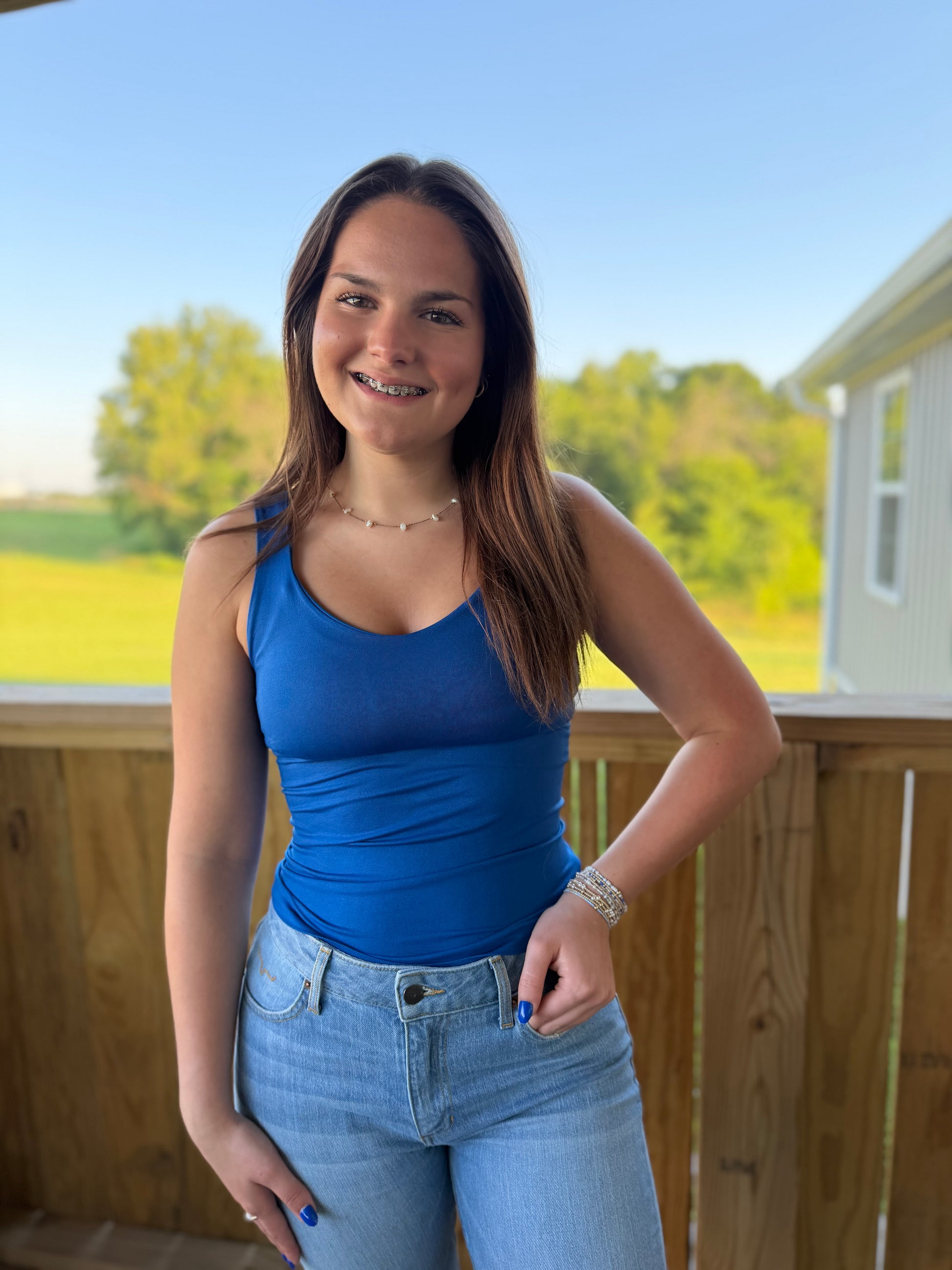 Woman in a blue tank top and jeans standing on a deck with a scenic background