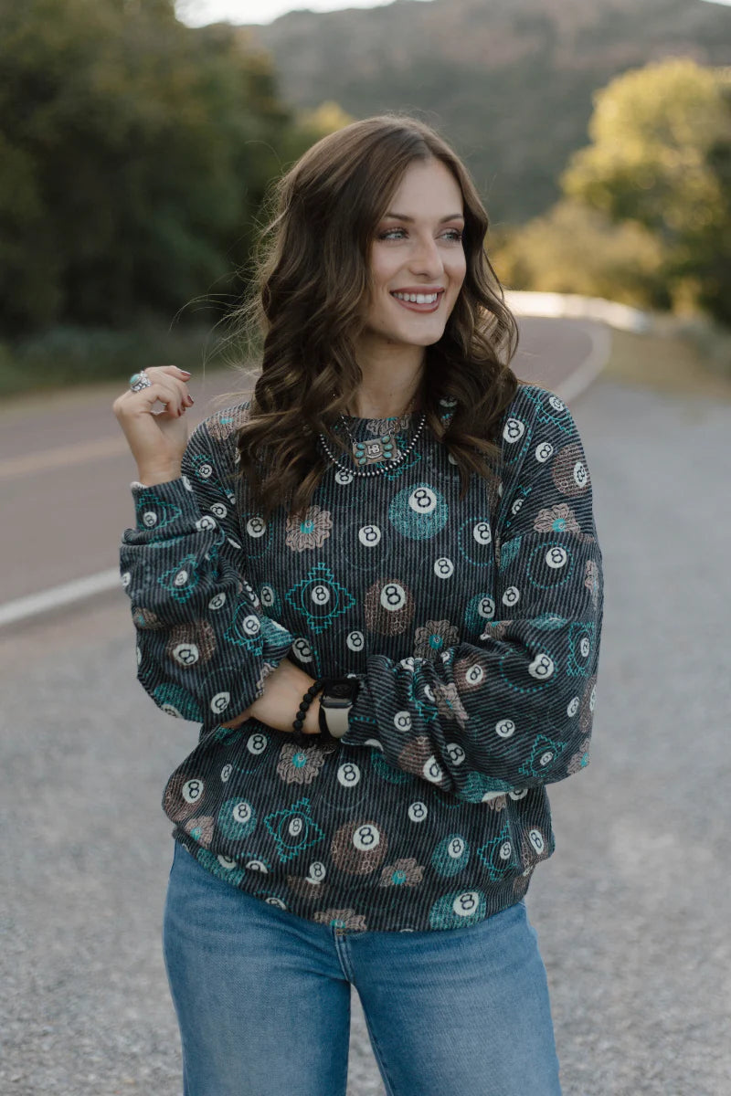 A woman wearing a long-sleeve sheer black top with a green and white circular floral pattern, paired with blue jeans, standing on an outdoor road.