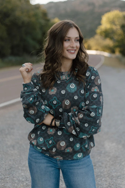 A woman wearing a long-sleeve sheer black top with a green and white circular floral pattern, paired with blue jeans, standing on an outdoor road.