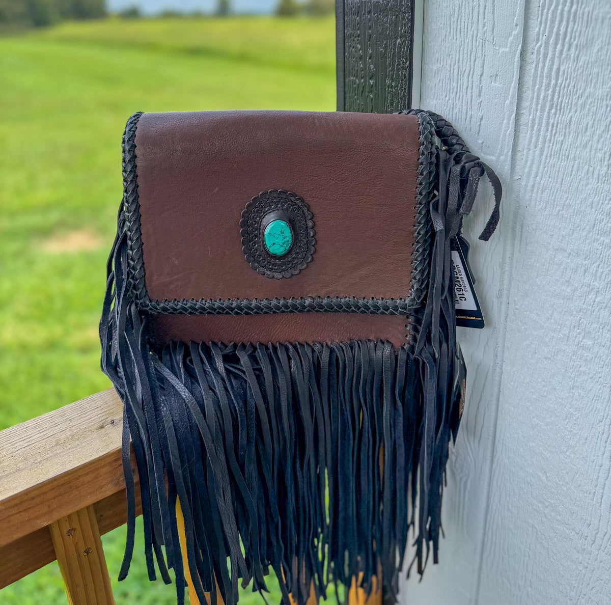 A brown leather crossbody bag with long black fringe and a turquoise stone accent on the front, photographed hanging against a white exterior wall with a blurred green field in the background.