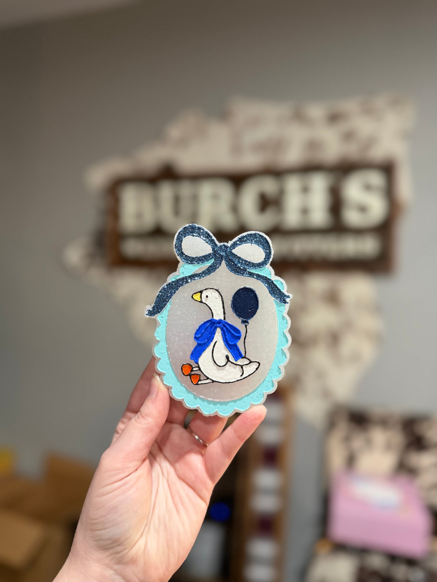 Hand holding a decorative item with a duck design in front of a 'Burch's' sign.