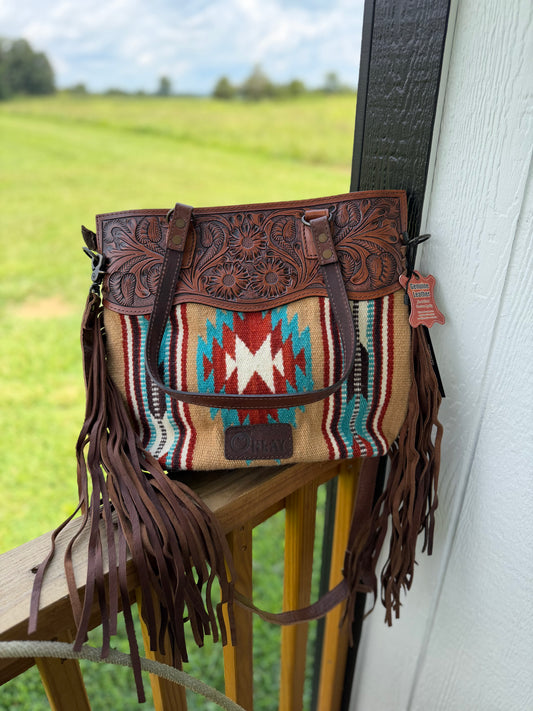 A western-style shoulder bag with brown tooled leather, a Southwestern patterned woven panel, and long brown fringe, resting on a wooden porch railing with a blurred green field and house siding in the background.