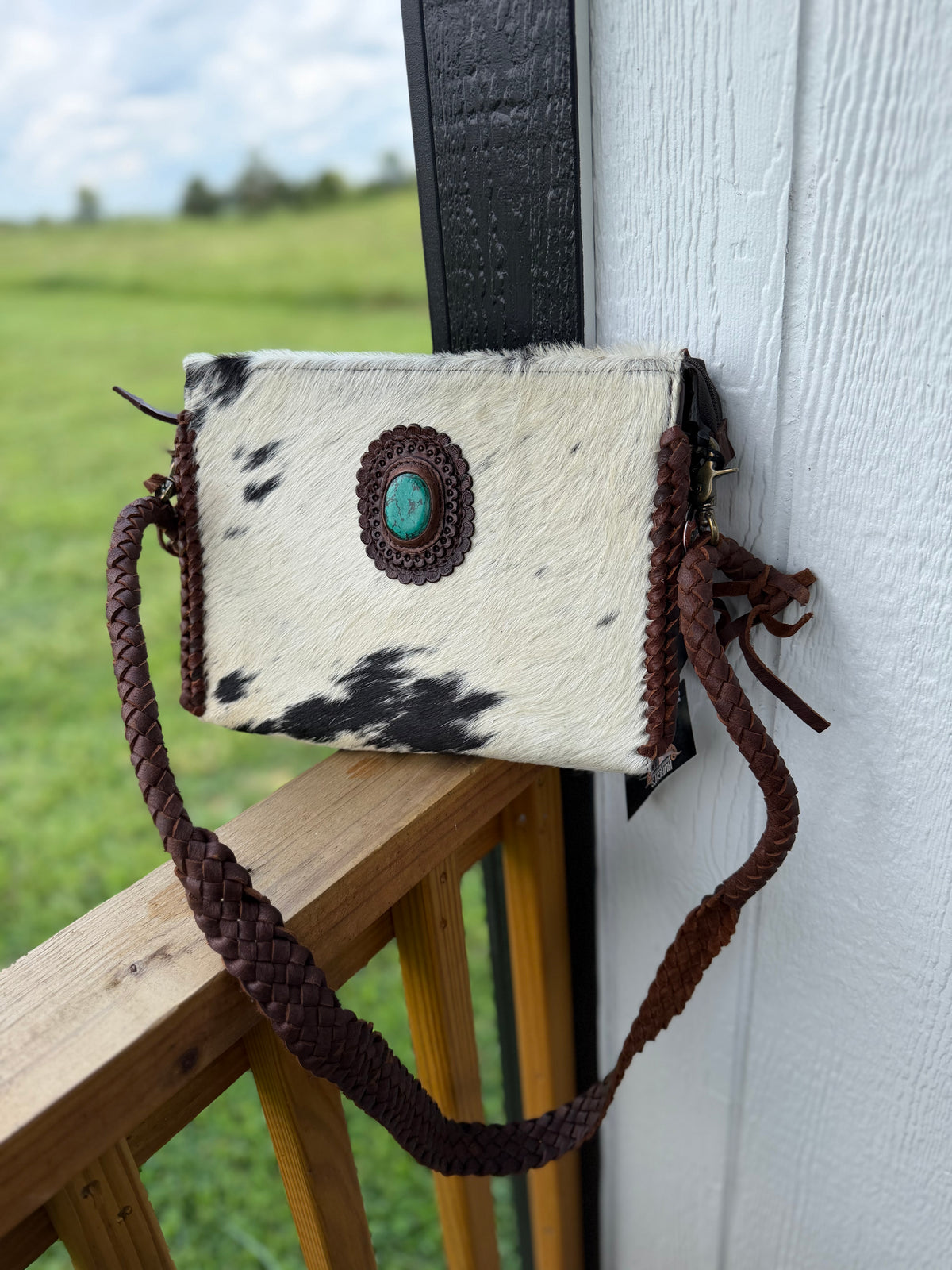A small cowhide crossbody bag with black and white spots, a brown braided strap, and a turquoise stone accent, hanging on a wooden railing next to a white wall.