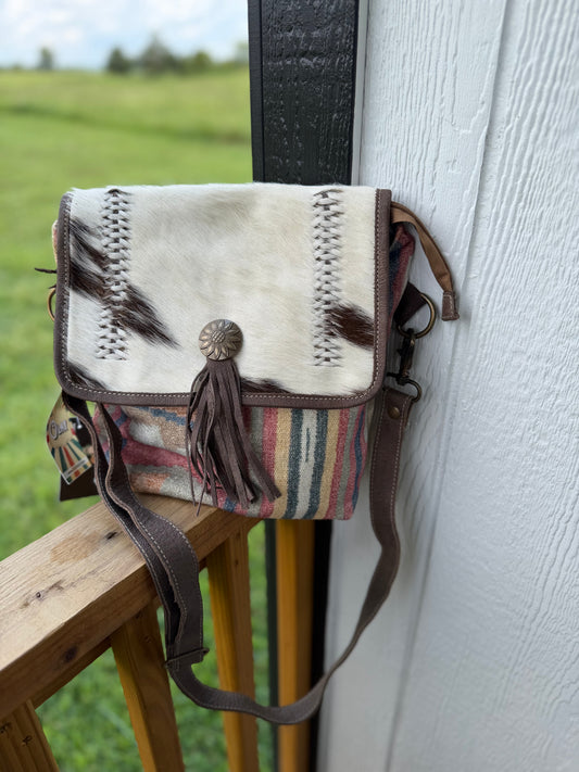 A western-style crossbody bag with a white cowhide flap, brown leather trim, a tassel, and a striped multicolor woven body, leaning against a white wooden wall next to a wooden railing with a blurred green field in the background.