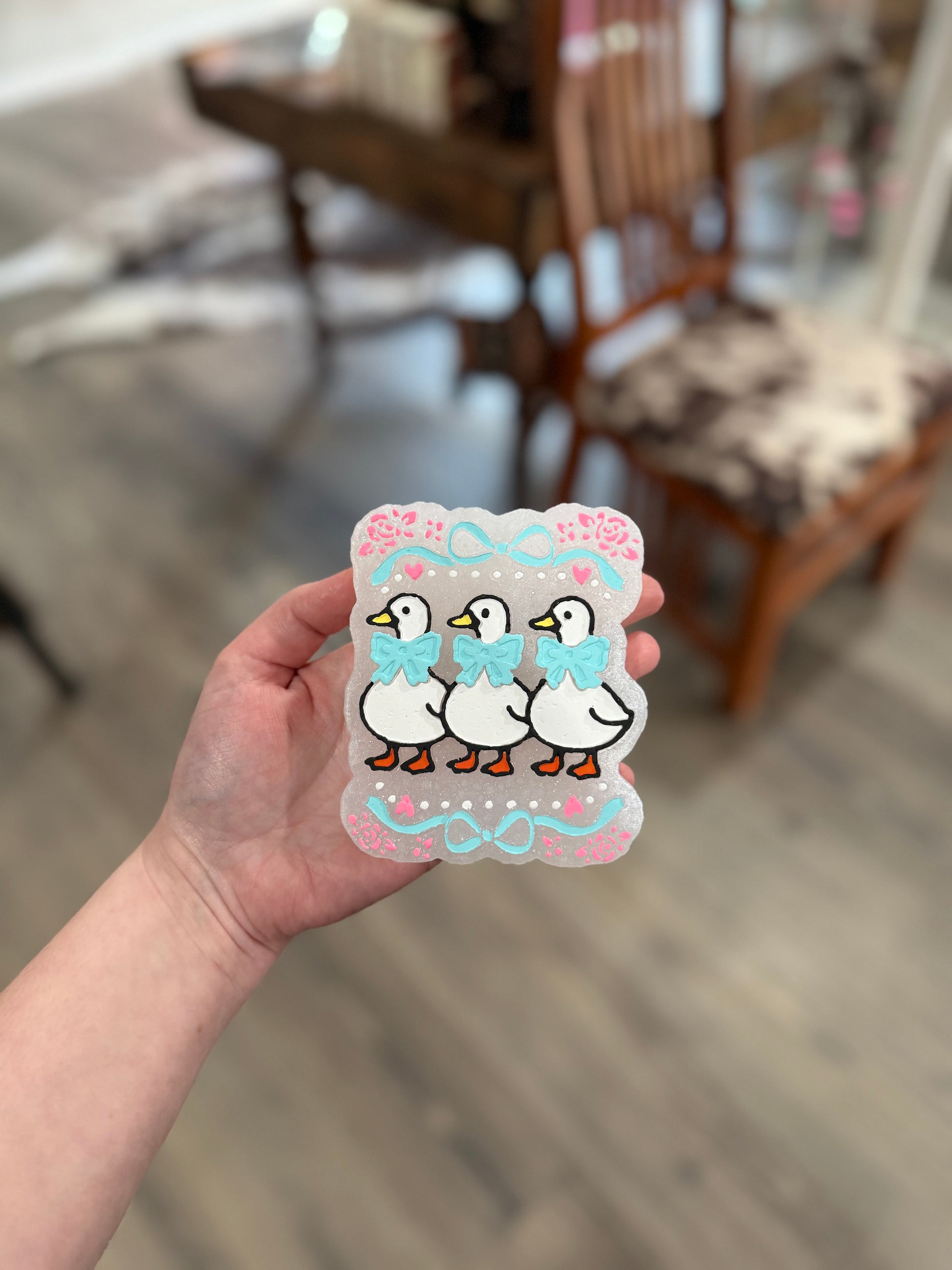 Hand holding a sticker with cartoon ducks in a room with wooden chairs and a rug.