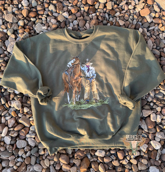 An olive green crewneck sweatshirt with a graphic print of a cowboy and his horse, laid flat on a bed of small brown and gray pebbles. A 'Purect's' brand logo is visible in the bottom right corner.