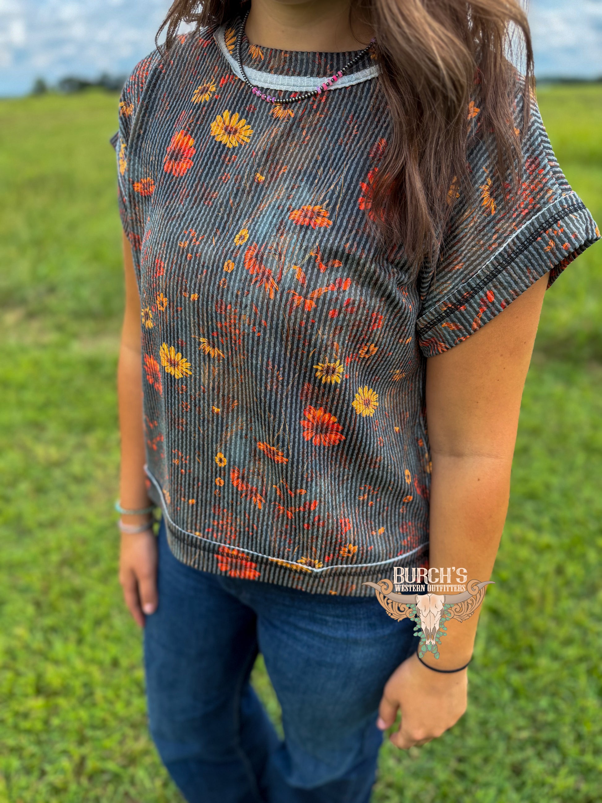 A woman wearing a short-sleeve ribbed knit top with a gray, orange, and yellow floral pattern, paired with blue jeans, standing in a grassy field.