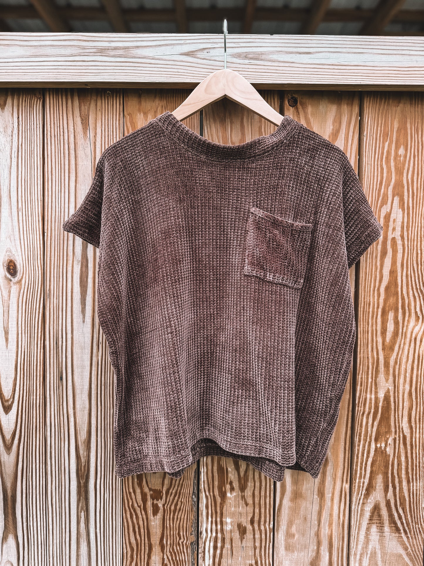 A short-sleeve waffle-knit t-shirt in a dark brownish-purple color, hanging on a wooden hanger against a rustic wooden plank wall.