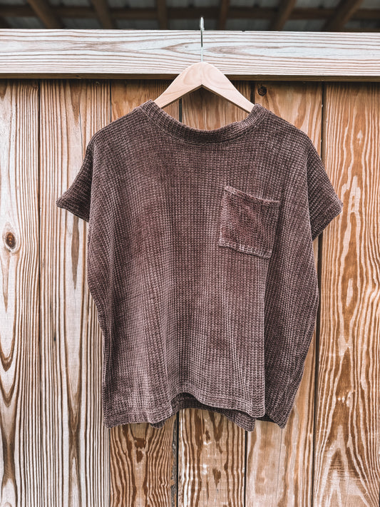 A short-sleeve waffle-knit t-shirt in a dark brownish-purple color, hanging on a wooden hanger against a rustic wooden plank wall.