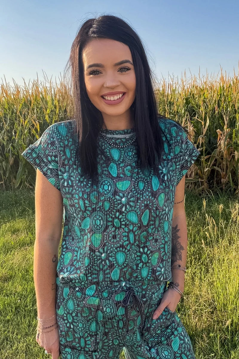 A person standing outdoors in a grassy field wearing loose, wide-leg pants with an allover teal, green, and dark gray paisley-style print, along with a matching top and sneakers.
