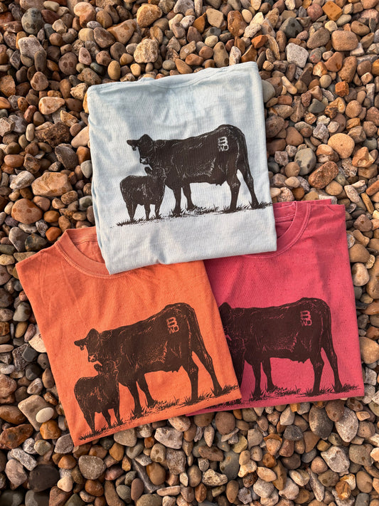 Three folded graphic T-shirts arranged on a bed of small brown pebbles. The top shirt is light blue with a black cow and calf silhouette; the bottom left is orange with a dark brown cow and calf silhouette; the bottom right is pink with a dark red cow and calf silhouette.