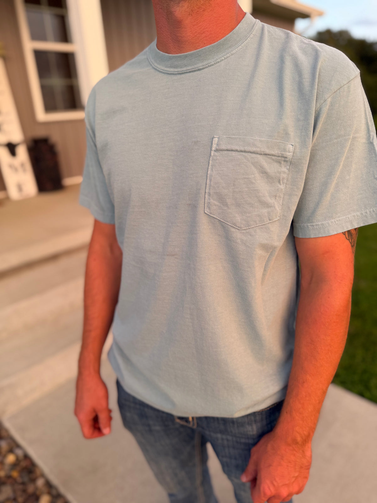 A man wearing a light blue crewneck pocket t-shirt and blue jeans, standing outdoors on a concrete porch in front of a house.