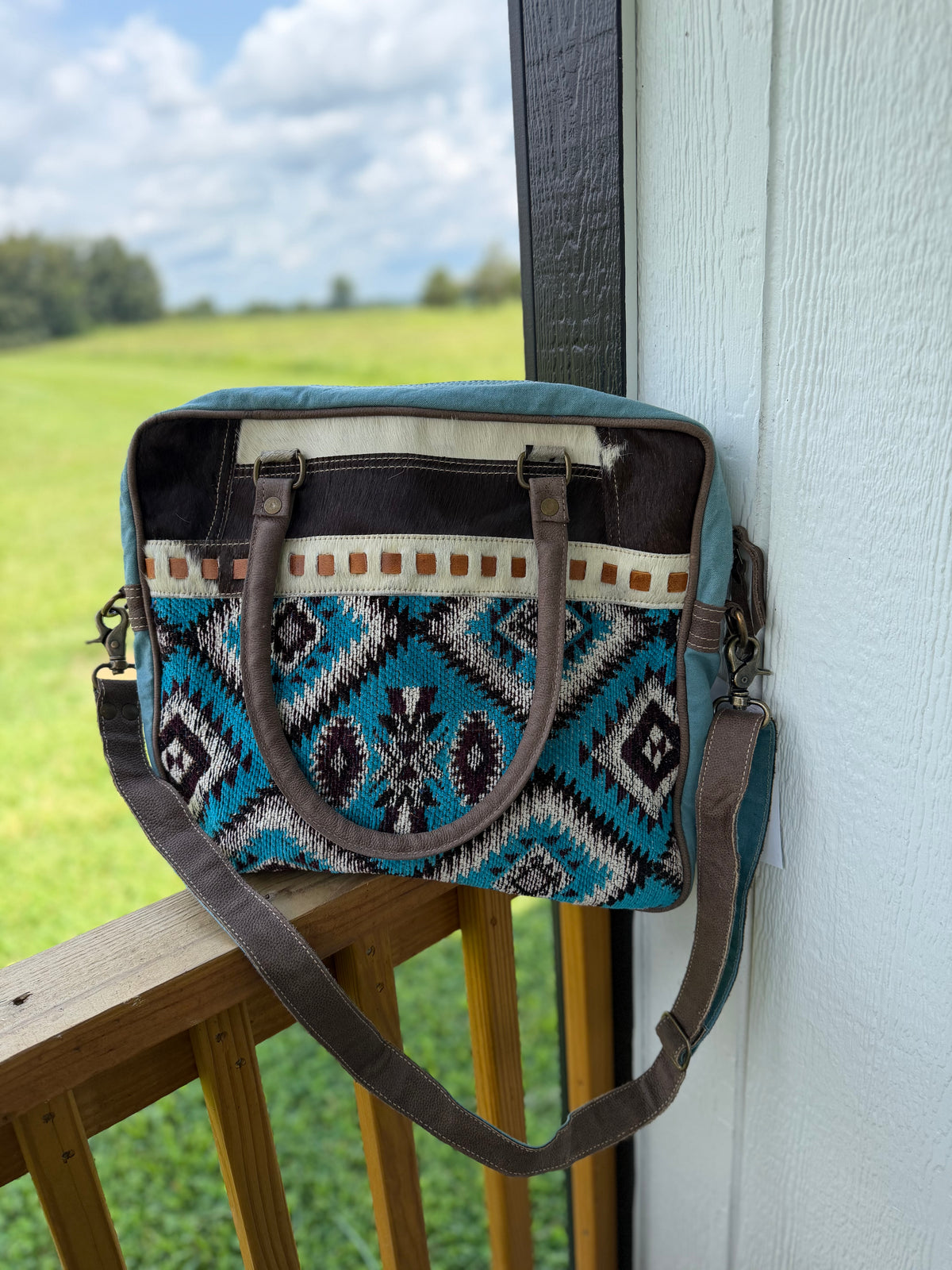 A woven shoulder bag with a Southwestern geometric pattern in turquoise, black, white, and brown, resting on a wooden deck railing next to a white wall with a green field in the background.