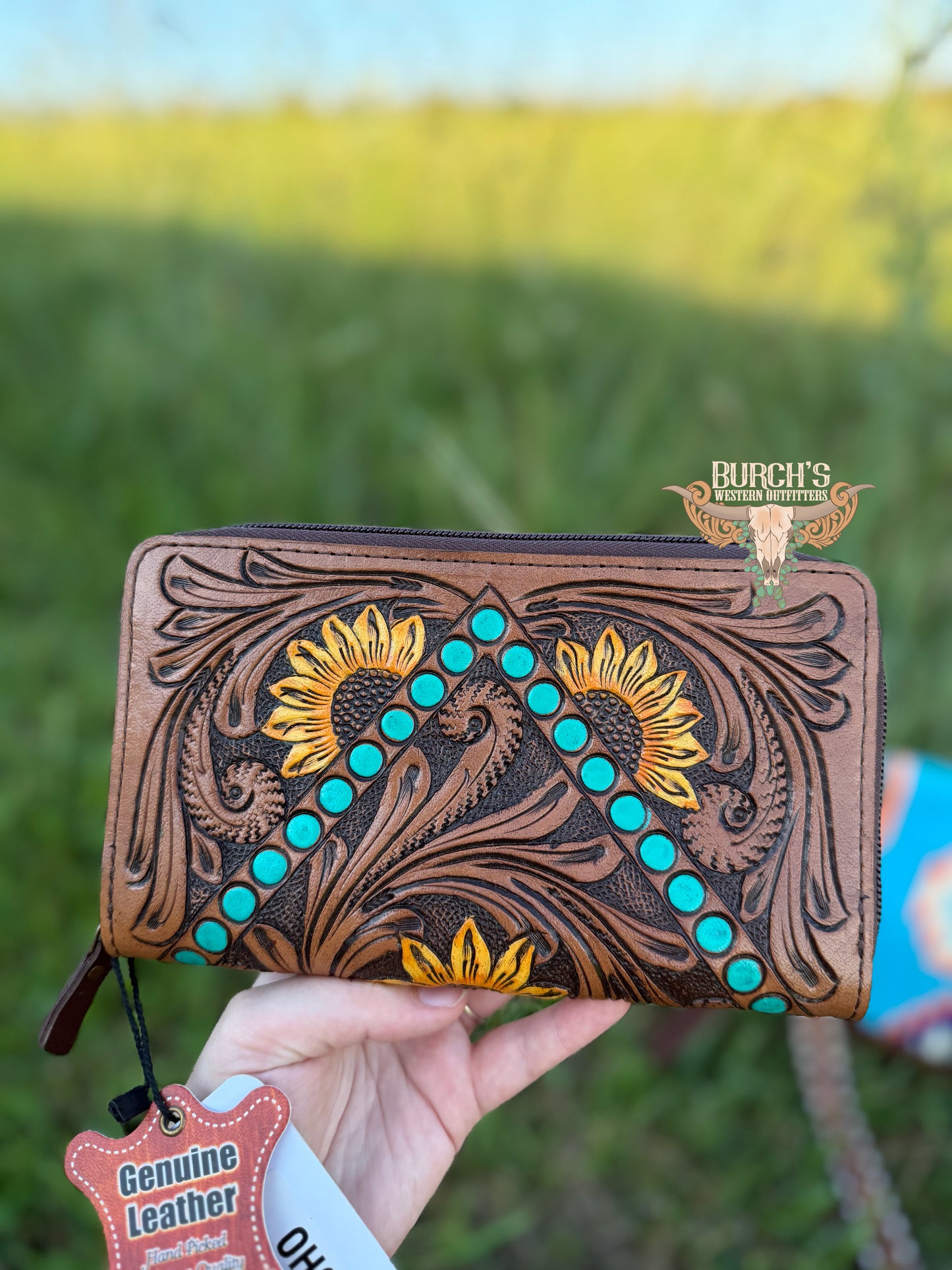 Handheld leather wallet with floral designs and turquoise stones against a blurred green field background.