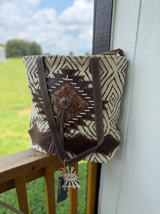 A woven bucket-style shoulder bag with a Southwestern geometric pattern in beige, brown, and white, featuring dark brown leather straps and a central tassel detail, hanging on a wooden railing next to a white wall.
