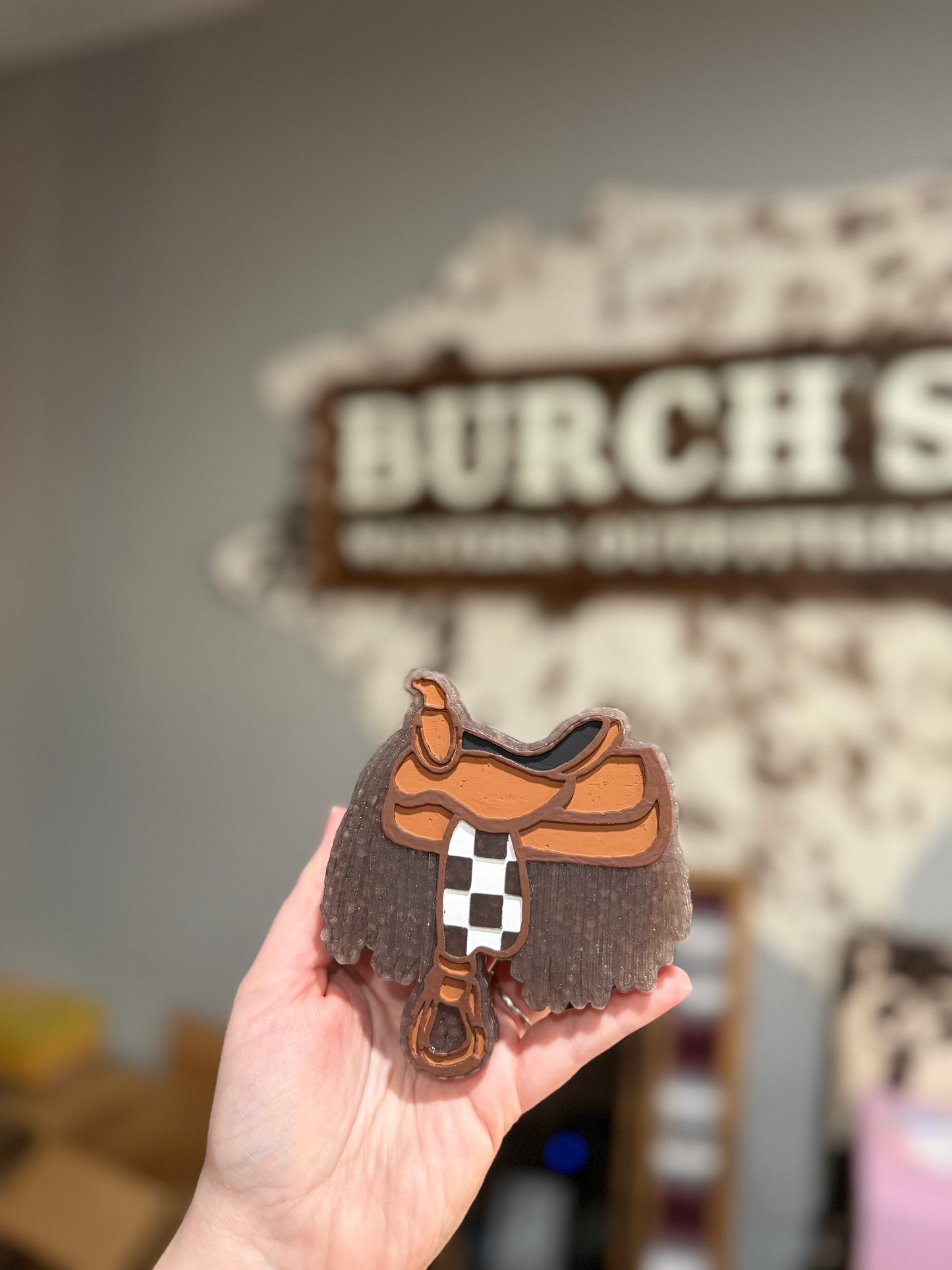 Hand holding a small leather saddle with a blurred 'Burch' sign in the background.