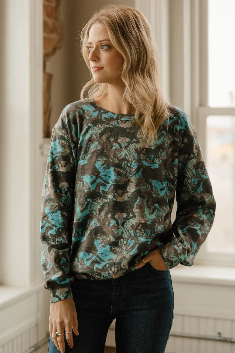 A woman wearing a long-sleeve crewneck sweatshirt with a dark green, brown, and teal abstract pattern, paired with dark blue jeans, standing in a brightly lit room.
