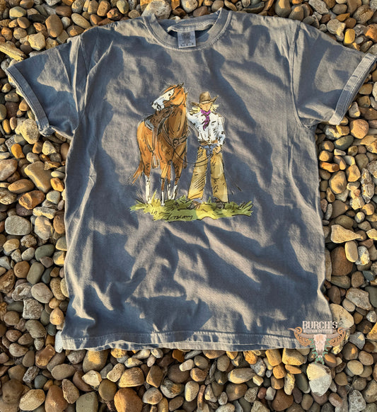 A faded blue-gray short-sleeve t-shirt with a western-themed graphic of a cowboy and a brown horse, laid flat on a bed of small brown and tan pebbles.