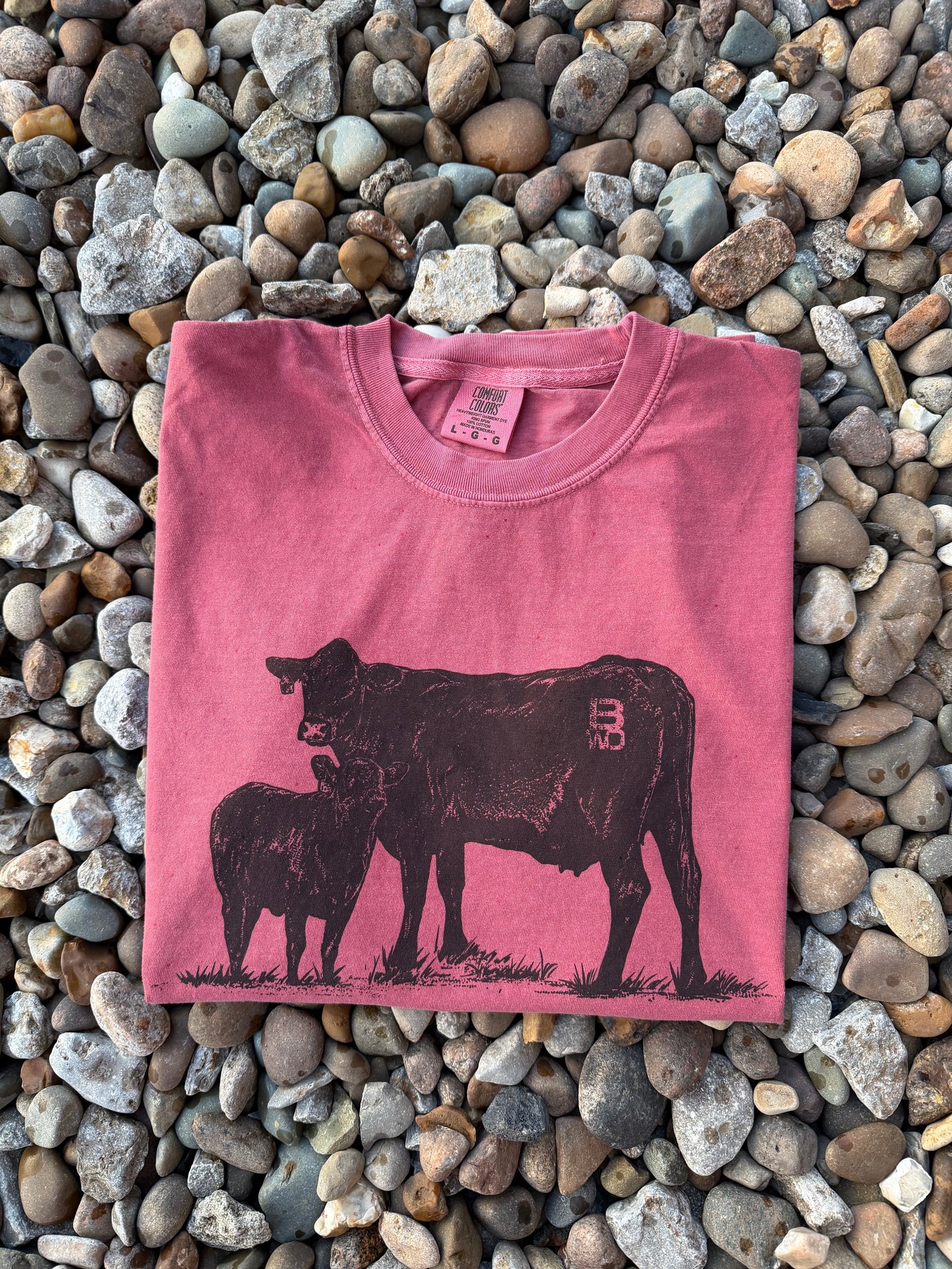 A folded pink t-shirt with a black cow and calf graphic, laid flat on a bed of small gray and brown pebbles. The shirt features a pink neck label with the brand name 'Buckeye'.