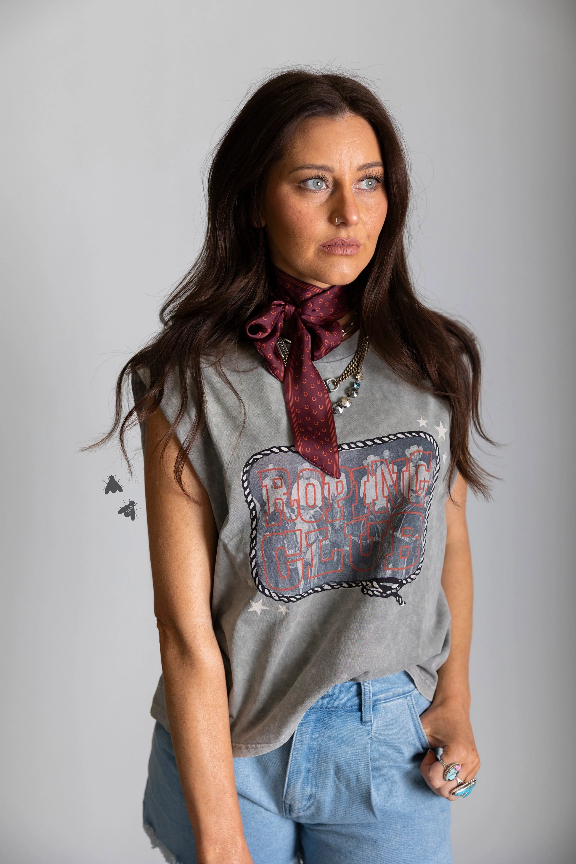 Woman wearing a sleeveless graphic shirt and blue jeans with a red scarf around her neck against a gray plain background