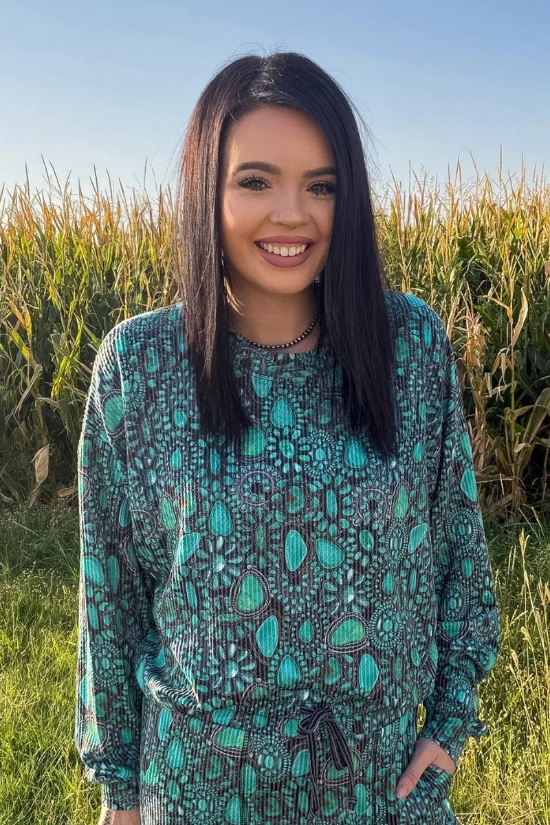 Woman modeling a turquoise and charcoal Western print long-sleeve top featuring a relaxed fit and turquoise stone-inspired pattern in front of a cornfield.