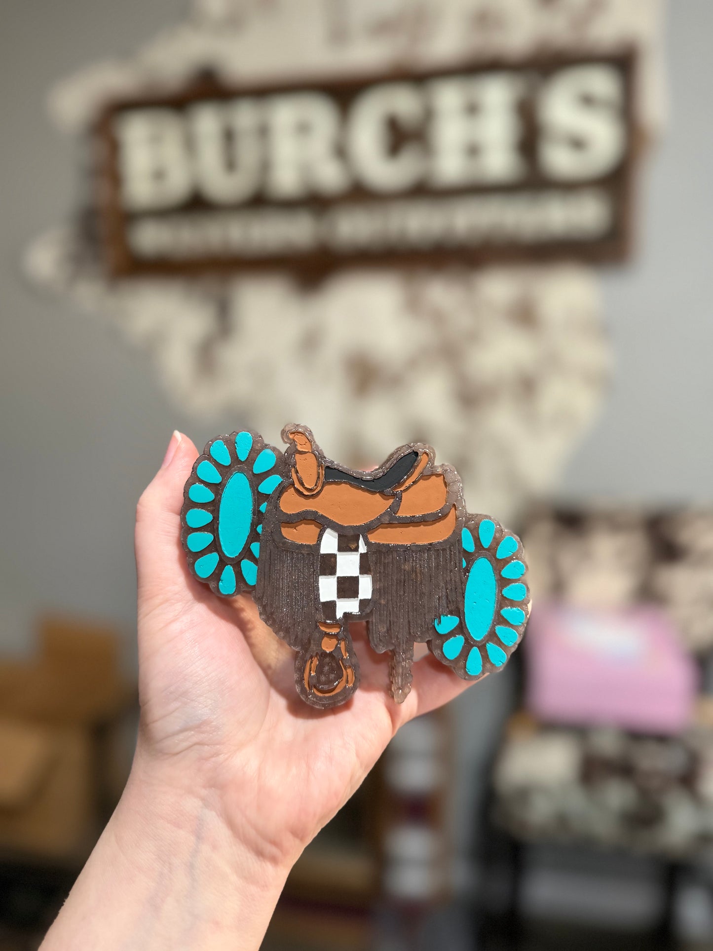 Hand holding a wooden boot with blue spurs and a checkered pattern, blurred background with 'Burch's' sign.