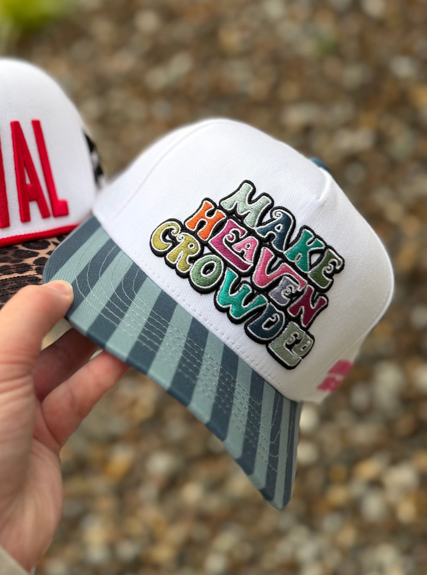 Two hats with text 'MAKE HEAVEN GROW' held by a hand against a blurred natural background.
