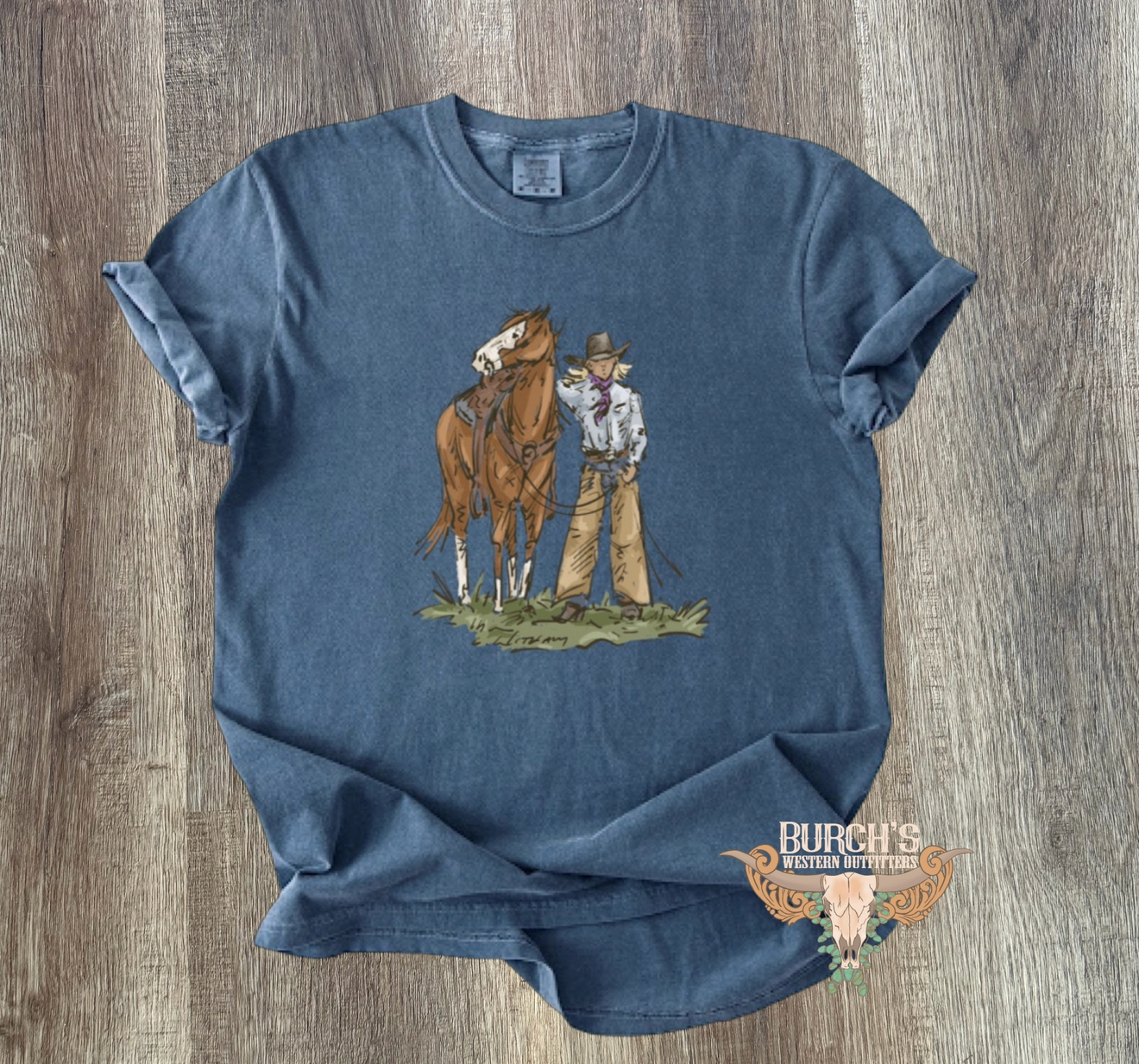 A blue short-sleeve graphic T-shirt laid flat on a wood surface, featuring a western-style illustration of a cowboy standing next to a horse and a small logo near the lower right hem.
