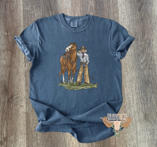 A blue short-sleeve graphic T-shirt laid flat on a wood surface, featuring a western-style illustration of a cowboy standing next to a horse and a small logo near the lower right hem.