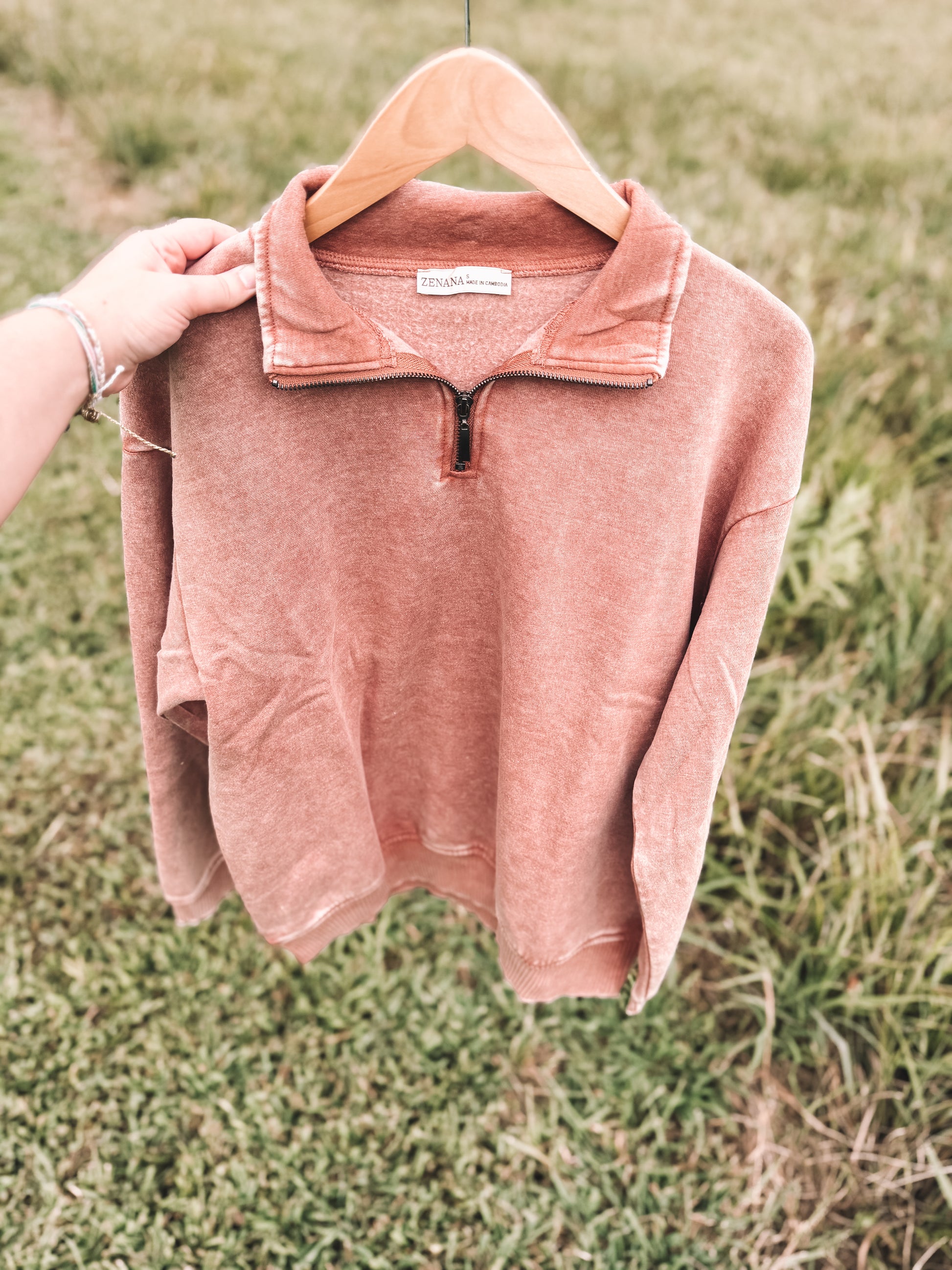 A dusty rose quarter-zip sweatshirt with a mock neck collar, hanging on a wooden hanger held by a person's hand against a blurred grassy background.