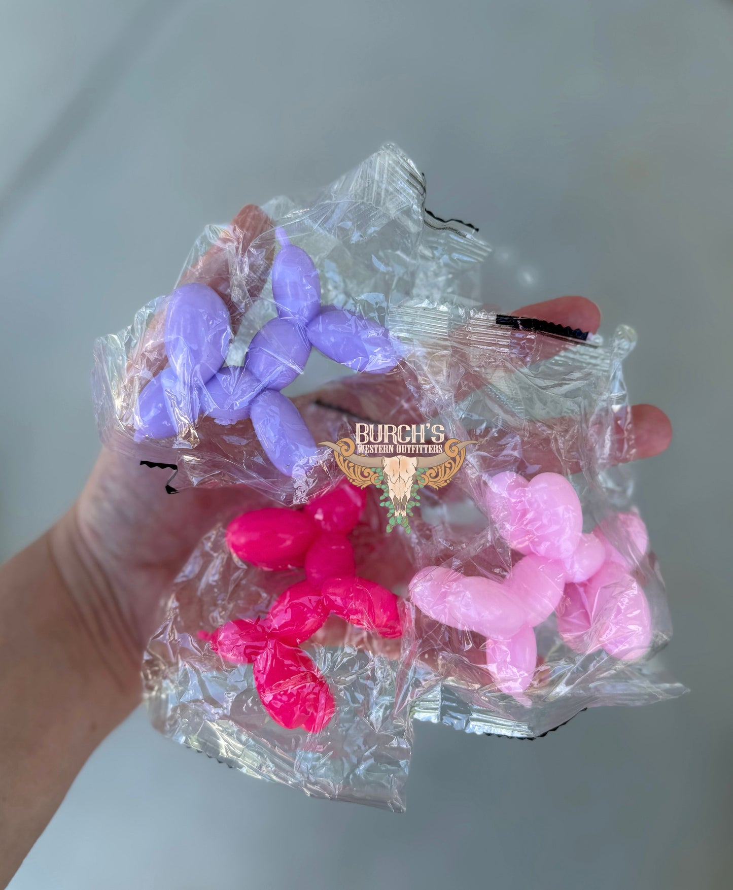 Pink and purple balloon animals in clear plastic packaging held by a hand on a light gray background.