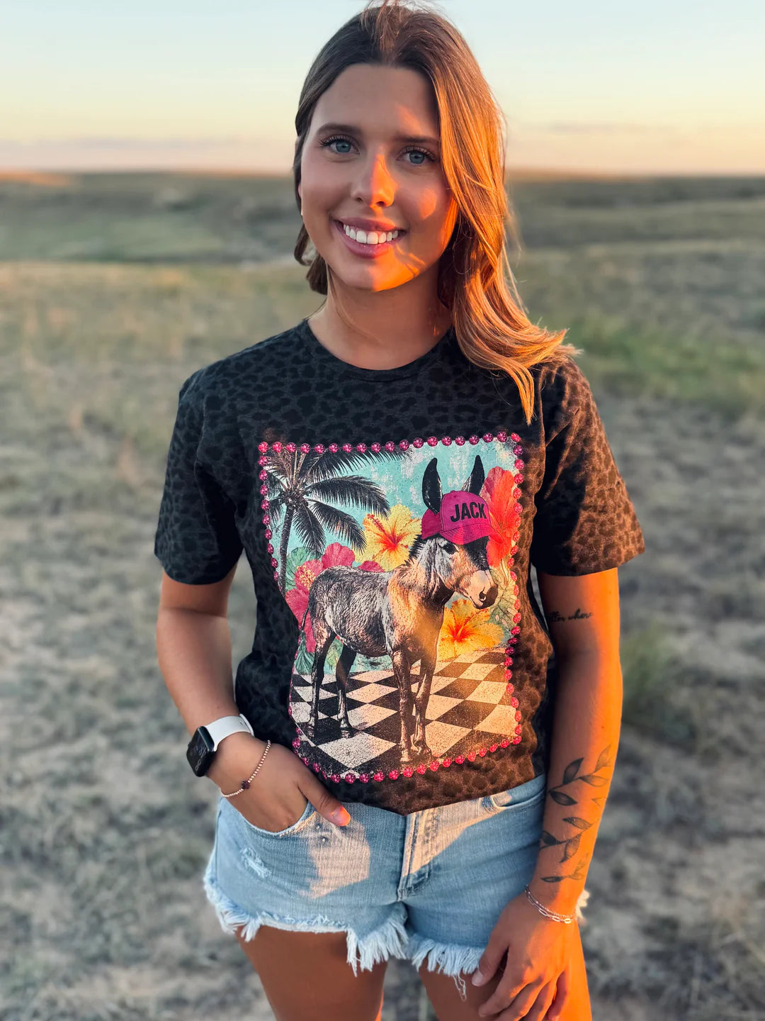 A woman wearing a black leopard-print graphic t-shirt with a colorful donkey illustration, paired with denim shorts, standing outdoors in a grassy field during sunset.