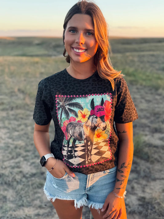 A woman wearing a black leopard-print graphic t-shirt with a colorful donkey illustration, paired with denim shorts, standing outdoors in a grassy field during sunset.