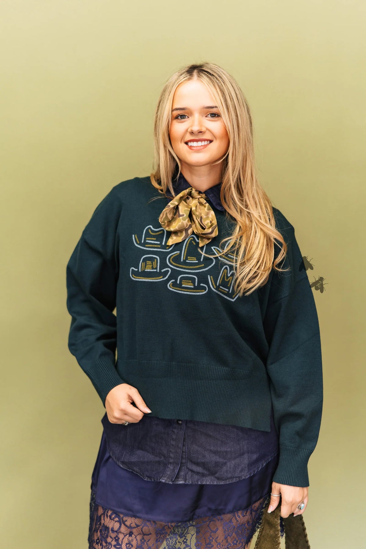 A smiling blonde woman wearing a dark teal-green crewneck sweatshirt with a graphic print, styled with a patterned neck scarf, a dark denim skirt, and lace socks, set against a solid olive-green studio background.