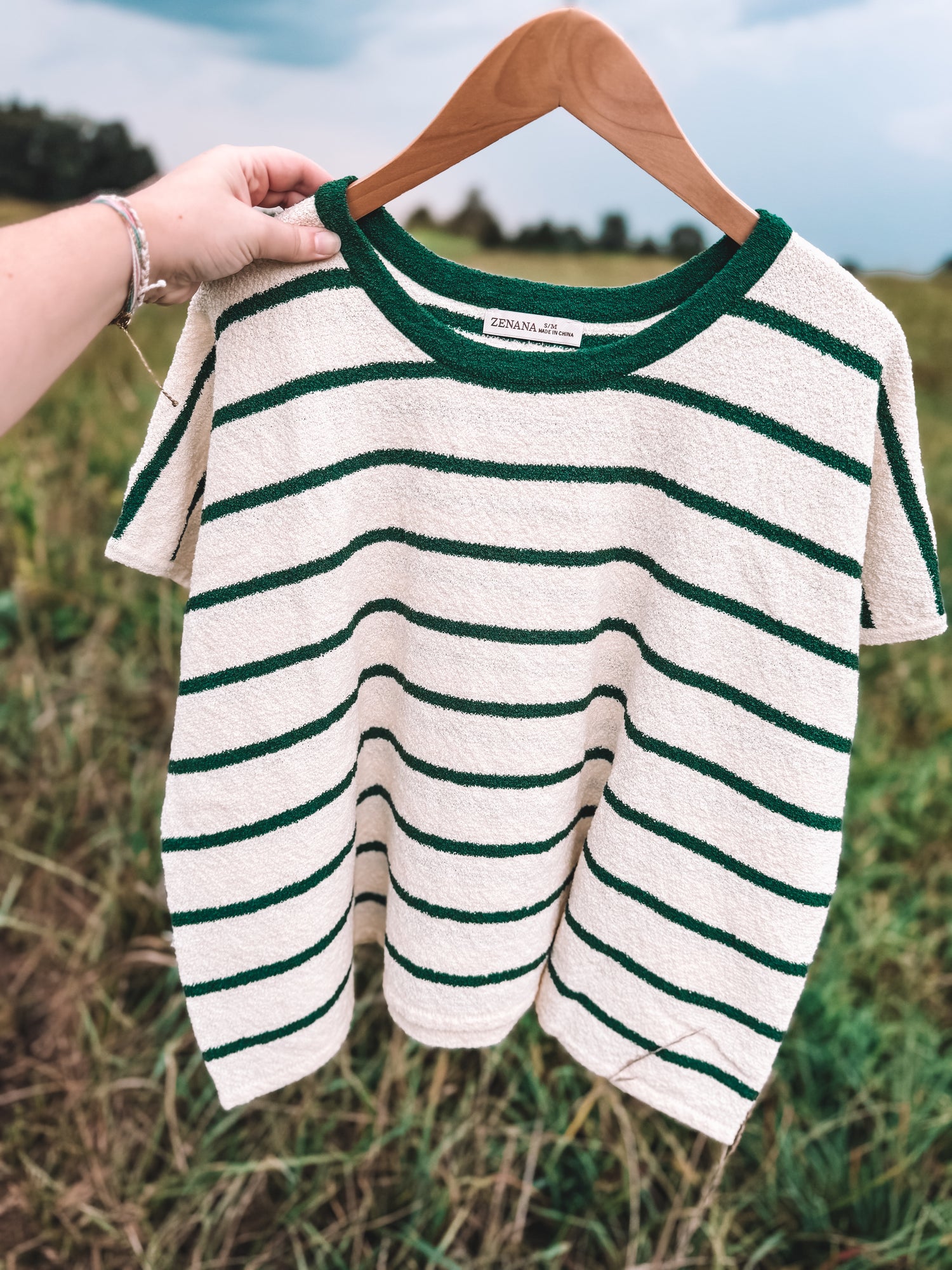 A short-sleeve knit top with dark green horizontal stripes on an off-white base, hanging on a wooden hanger held by a person's hand against an outdoor grassy background.